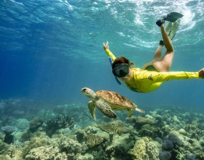 A snorkeler in a yellow wetsuit swims alongside a sea turtle over a vibrant coral reef. The scene conveys a sense of serenity and connection with nature.