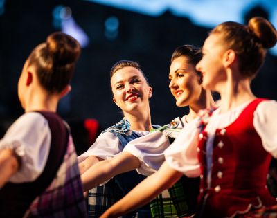 Four smiling women perform a traditional Scottish Highland dance, wearing colorful tartan dresses, with a building visible in the background.