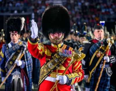 A member of a traditional Scottish military band stands at attention, facing forward, in front of other band members and a muted audience.