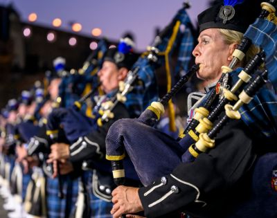 A line of bagpipers in traditional Scottish uniforms, the foremost a blonde woman playing with intense concentration, illuminated against a soft twilight backdrop.