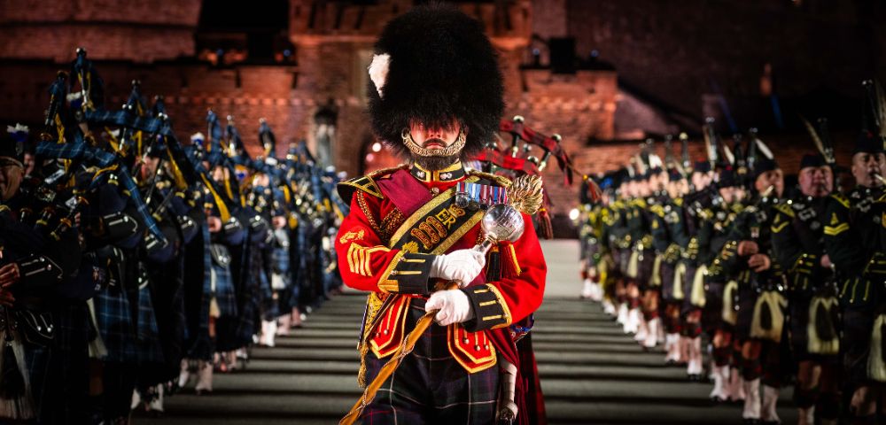 A member of a Scottish military band in full regalia, including a tall, feathered hat and red military coat, stands at attention in the foreground, with rows of other band members stretching into the background in formation.