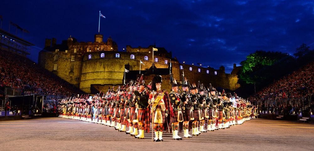 A large group of uniformed bagpipers in traditional Scottish dress, lined up in rows with Edinburgh Castle illuminated in the background, face towards the foreground, while spectators fill the stands.