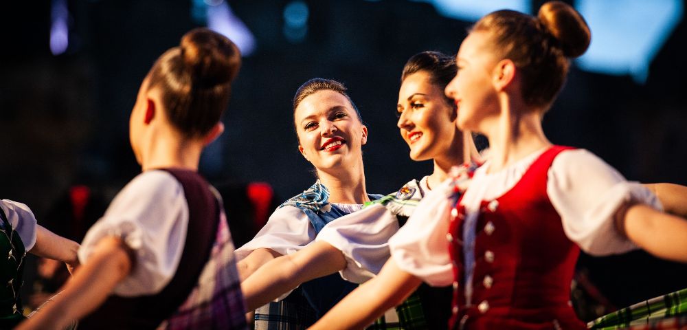 Four smiling women perform a traditional Scottish Highland dance, wearing colorful tartan dresses, with a building visible in the background.