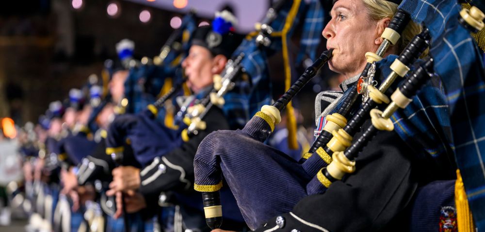A line of bagpipers in traditional Scottish uniforms, the foremost a blonde woman playing with intense concentration, illuminated against a soft twilight backdrop.