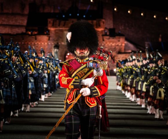 A member of a Scottish military band in full regalia, including a tall feathered hat and red military coat, stands at attention in the foreground, with rows of other band members stretching into the background in formation.