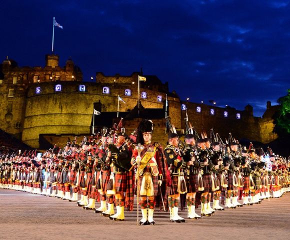 A large group of uniformed bagpipers in traditional Scottish dress, lined up in rows with Edinburgh Castle illuminated in the background, face towards the foreground, while spectators fill the stands.