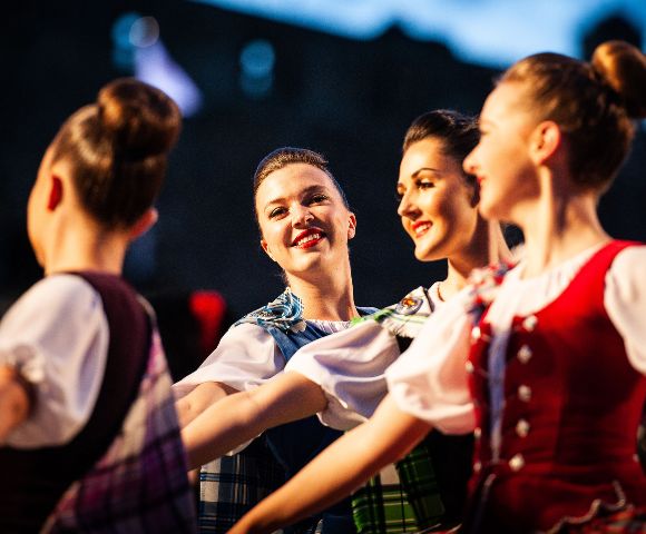 Four smiling women perform a traditional Scottish Highland dance, wearing colorful tartan dresses, with a building visible in the background.