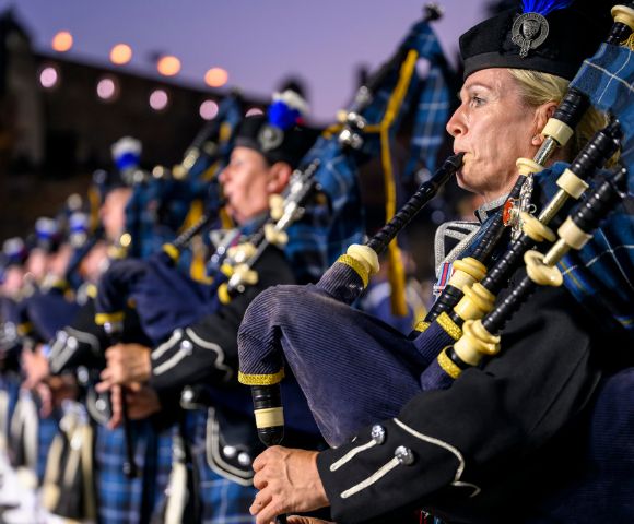 A line of bagpipers in traditional Scottish uniforms, the foremost a blonde woman playing with intense concentration, illuminated against a soft twilight backdrop.