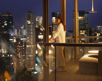 A woman in a white outfit stands on a balcony at night, gazing at a city skyline with illuminated buildings. The scene conveys a serene, contemplative mood.