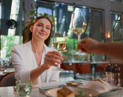 A joyful woman in a chic white blazer toasts with a glass of wine in an elegant restaurant setting.