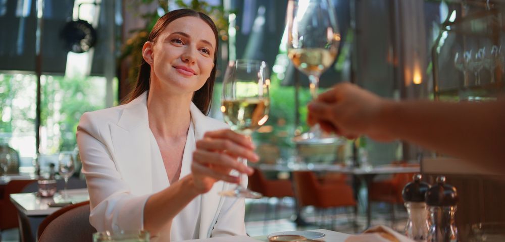 A joyful woman in a chic white blazer toasts with a glass of wine in an elegant restaurant setting.