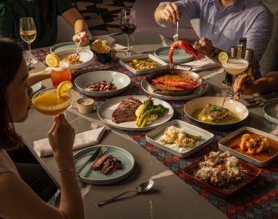 A group enjoys a vibrant meal at a dining table. Dishes include paella, steak, and shrimp, with cocktails and wine. The scene is warm and inviting.
