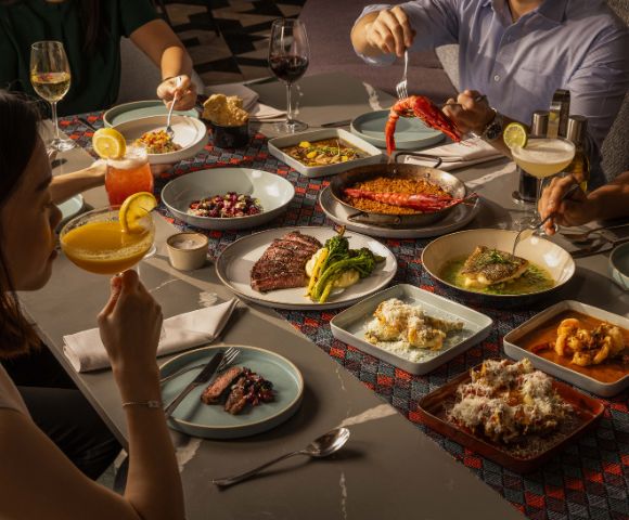 A group enjoys a vibrant meal at a dining table. Dishes include paella, steak, and shrimp, with cocktails and wine. The scene is warm and inviting.