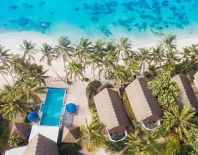Aerial view of a tropical resort with thatched-roof huts, a swimming pool, and palm trees beside a turquoise beach.