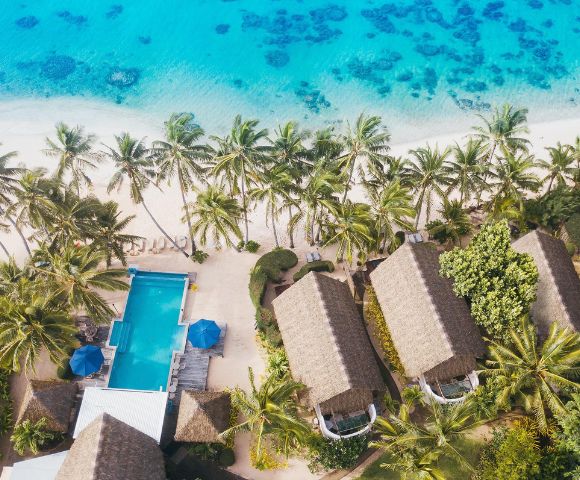 Aerial view of a tropical resort with thatched-roof huts, a swimming pool, and palm trees beside a turquoise beach.