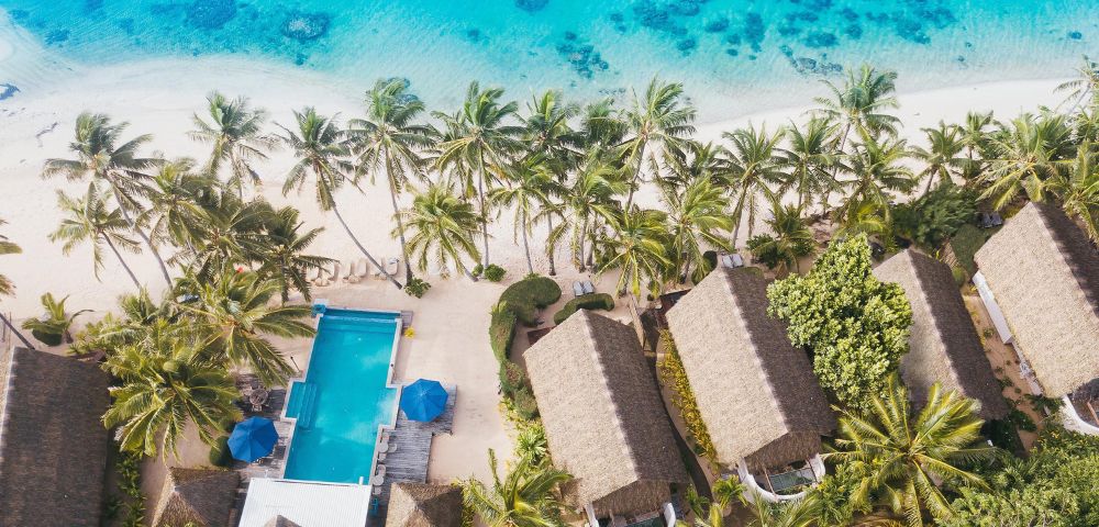 Aerial view of a tropical resort with thatched-roof huts, a swimming pool, and palm trees beside a turquoise beach.