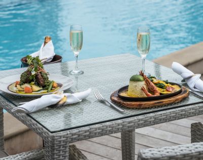 Two elegant plates of gourmet dishes are set on a glass table beside a blue pool, accompanied by flutes of sparkling wine.