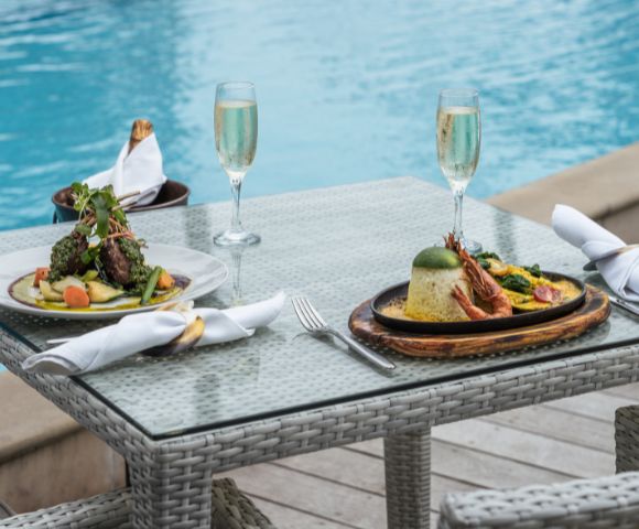 Two elegant plates of gourmet dishes are set on a glass table beside a blue pool, accompanied by flutes of sparkling wine.
