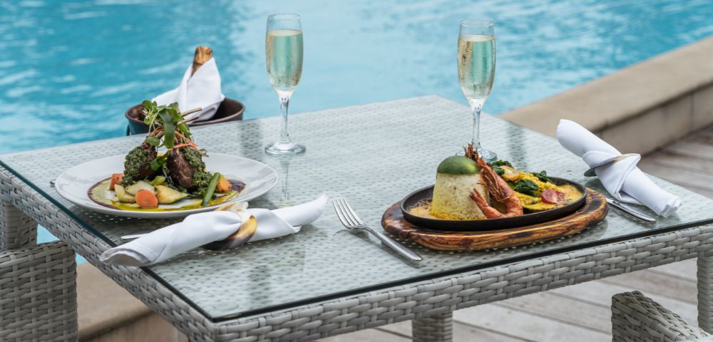 Two elegant plates of gourmet dishes are set on a glass table beside a blue pool, accompanied by flutes of sparkling wine.