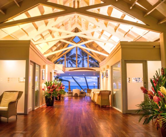 Cozy interior of a tropical restaurant featuring a high ceiling, wooden floors, and tropical plants, with an ocean view.