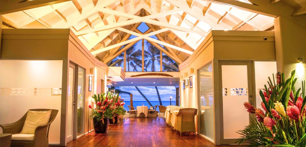 Cozy interior of a tropical restaurant featuring a high ceiling, wooden floors, and tropical plants, with an ocean view.