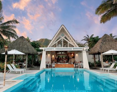 Tropical poolside scene featuring a modern restaurant with a peaked roof, surrounded by palm trees and guests enjoying the view at sunset.