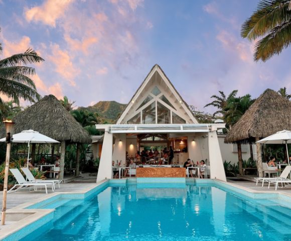 Tropical poolside scene featuring a modern restaurant with a peaked roof, surrounded by palm trees and guests enjoying the view at sunset.