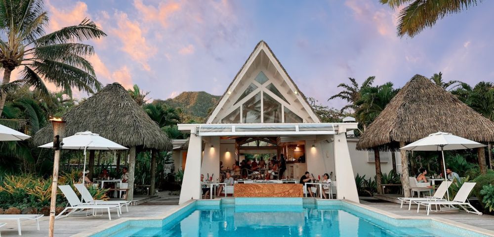 Tropical poolside scene featuring a modern restaurant with a peaked roof, surrounded by palm trees and guests enjoying the view at sunset.