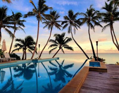 A serene tropical scene with tall palm trees framing an infinity pool overlooking a calm ocean, set against a vibrant sunset sky. Relaxing and tranquil atmosphere.