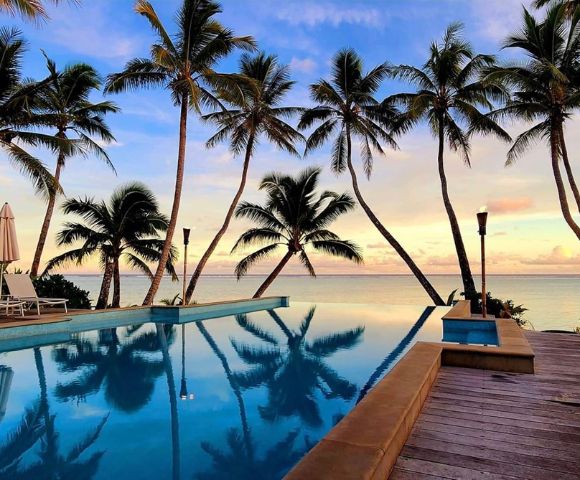 A serene tropical scene with tall palm trees framing an infinity pool overlooking a calm ocean, set against a vibrant sunset sky. Relaxing and tranquil atmosphere.