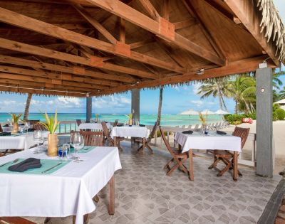 Outdoor beachside restaurant under a wooden canopy, with tables set for dining. Ocean and palm trees in the background, conveying a relaxed, tropical vibe.