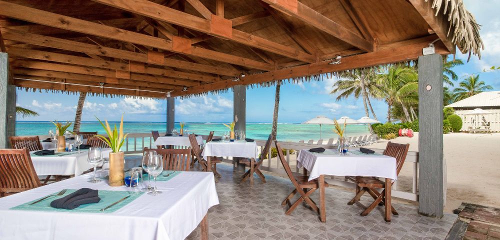 Outdoor beachside restaurant under a wooden canopy, with tables set for dining. Ocean and palm trees in the background, conveying a relaxed, tropical vibe.