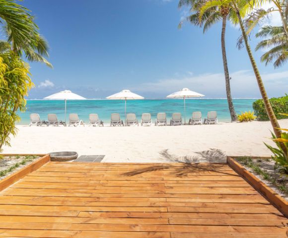 Tropical beach scene with wooden walkway leading to white sand, rows of sun loungers and umbrellas, surrounded by palm trees, under a bright blue sky.