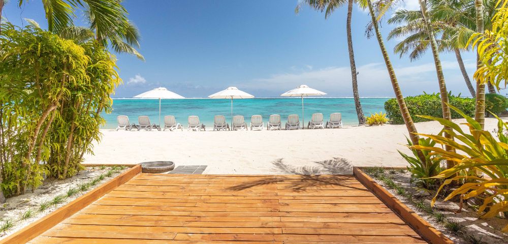 Tropical beach scene with wooden walkway leading to white sand, rows of sun loungers and umbrellas, surrounded by palm trees, under a bright blue sky.