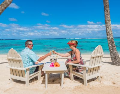 Couple toasting champagne glasses on a sandy tropical beach. They sit in chairs under palm trees, facing turquoise ocean under a bright blue sky. Relaxing mood.