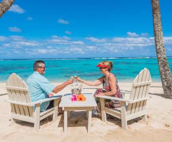 Couple toasting champagne glasses on a sandy tropical beach. They sit in chairs under palm trees, facing turquoise ocean under a bright blue sky. Relaxing mood.