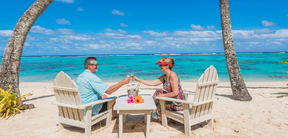 Couple toasting champagne glasses on a sandy tropical beach. They sit in chairs under palm trees, facing turquoise ocean under a bright blue sky. Relaxing mood.