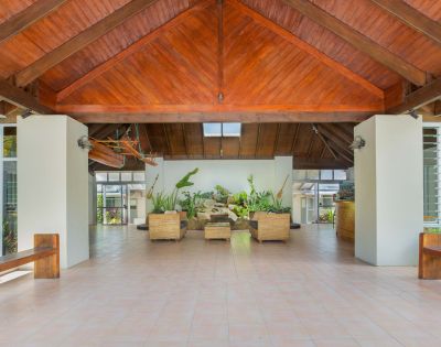 Spacious, open-air atrium with a wooden, vaulted ceiling and tiled floor. Wicker seating, lush plants, and natural light create a serene, inviting atmosphere