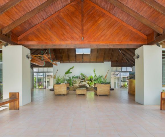 Spacious, open-air atrium with a wooden, vaulted ceiling and tiled floor. Wicker seating, lush plants, and natural light create a serene, inviting atmosphere