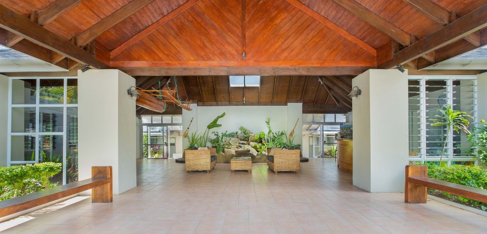 Spacious, open-air atrium with a wooden, vaulted ceiling and tiled floor. Wicker seating, lush plants, and natural light create a serene, inviting atmosphere