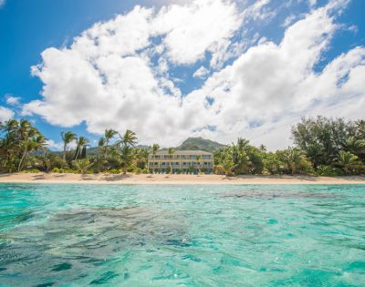 Sunny beach scene with clear turquoise water in the foreground, palm trees lining a sandy shore, and a bright sun and fluffy clouds in a blue sky.