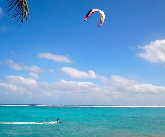 Kitesurfer rides on turquoise ocean under a vibrant blue sky with scattered clouds, feeling adventurous. A palm leaf peeks from the top left corner.