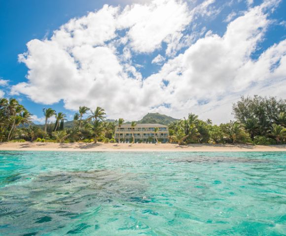 Sunny beach scene with clear turquoise water in the foreground, palm trees lining a sandy shore, and a bright sun and fluffy clouds in a blue sky.