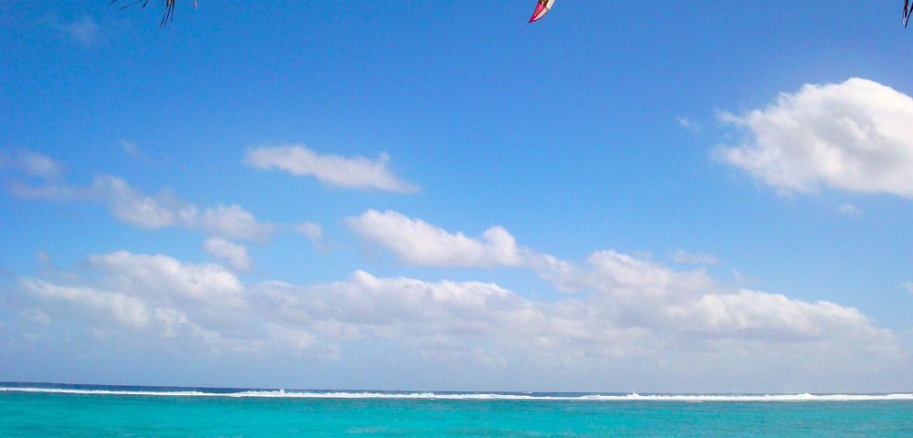 Kitesurfer rides on turquoise ocean under a vibrant blue sky with scattered clouds, feeling adventurous. A palm leaf peeks from the top left corner.