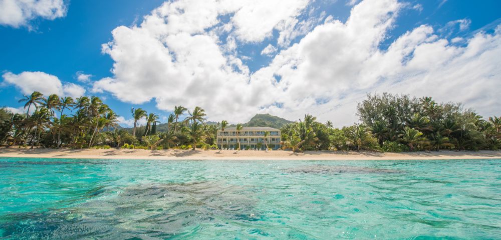 Sunny beach scene with clear turquoise water in the foreground, palm trees lining a sandy shore, and a bright sun and fluffy clouds in a blue sky.