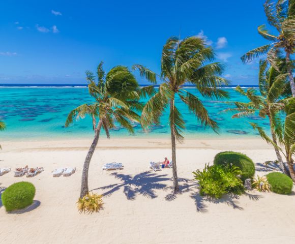 Sunny beach scene with turquoise water, white sand, and swaying palm trees. People relax on lounge chairs, conveying a peaceful, tropical vibe.