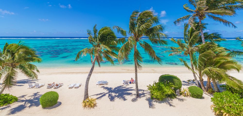 Sunny beach scene with turquoise water, white sand, and swaying palm trees. People relax on lounge chairs, conveying a peaceful, tropical vibe.