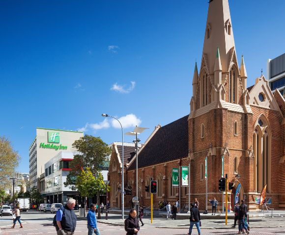 A sunny street scene with people walking past a historic brick church with a tall spire. Modern buildings and a Holiday Inn are visible in the background.