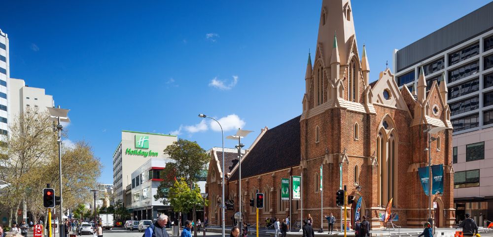 A sunny street scene with people walking past a historic brick church with a tall spire. Modern buildings and a Holiday Inn are visible in the background.