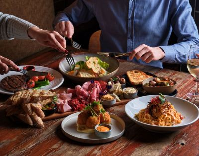 A wooden table with a shared meal featuring steak, salad, pasta, cold cuts, bread, and olives. Three hands reach in, creating a communal, inviting atmosphere.